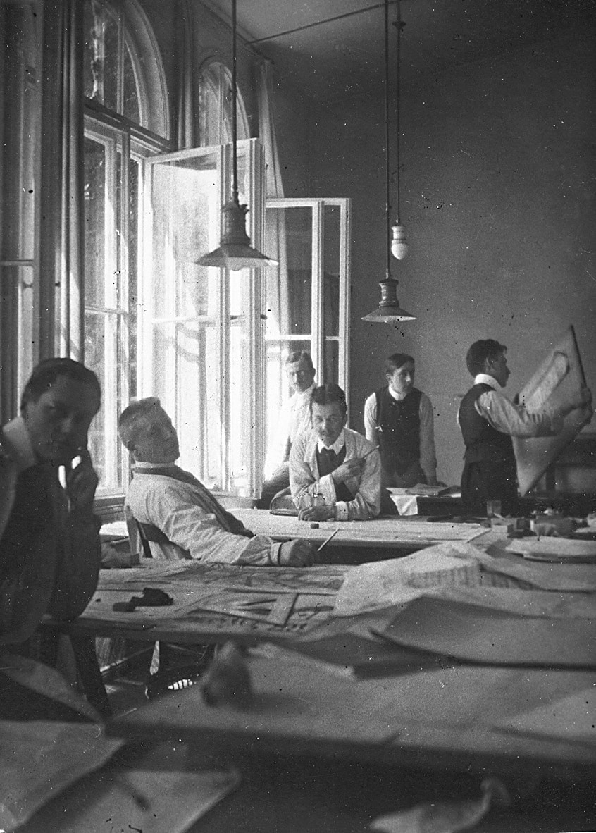 Sechs Assistenten von Behrens am Arbeitsplatz: (von li.) Mies van der Rohe, Meyer, Hertwig, Weyrather (dahinter), Krämer, Gropius (mit Plan), 1908[